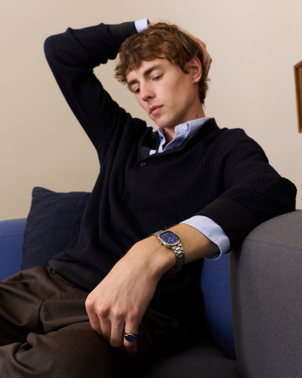 Seorang pria mengenakan jam tangan dengan tali logam dan dial biru duduk di sofa