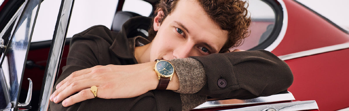 Seorang pria mengenakan jam tangan Daniel Wellington dengan tali kulit cokelat dan dial hijau di pergelangan tangan kirinya sambil bersandar pada mobil merah